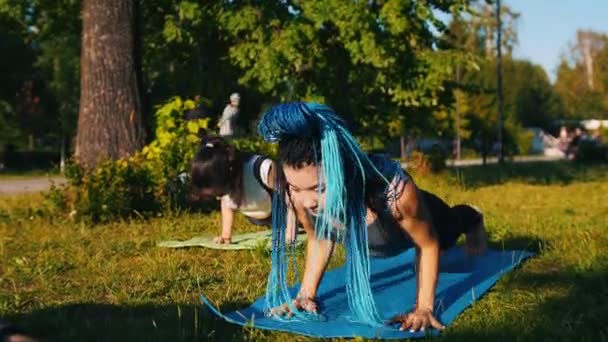 Две молодые атлетичные женщины, занимающиеся йогой в парке - Одна женщина имеет длинные синие дреды — стоковое видео
