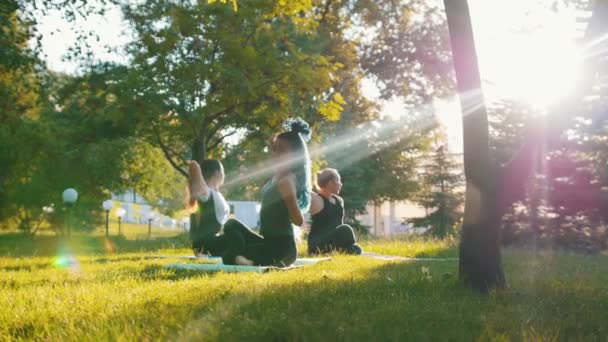 Två unga kvinnor gör yoga övningar med tränare i parken i solens strålar-en kvinna har långa blå dreadlocks — Stockvideo