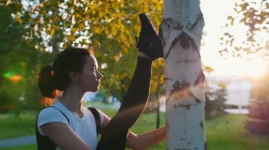 Genç çekici anne güneş ışınları parkta ağacın yanında germe yapıyor - Raising ve ağaca karşı bacağını eğilerek