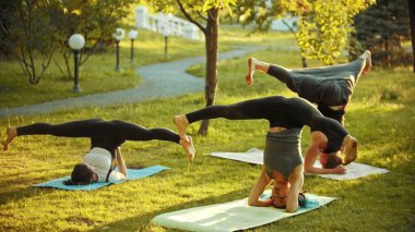 Parlak yeşil ağaçlar ve geçit yakınındaki parkta bir eğitmen ile yoga egzersizleri yapan iki genç kadın - Headstands doing