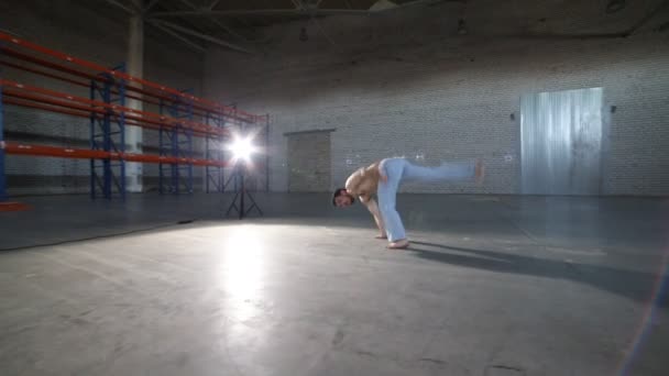 Un homme athlétique faisant des éléments de capoeira difficiles dans la pièce avec sol en béton et murs en briques — Video