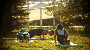 İki genç kadın ve bir adam parkta yoga yapıyor parlak yeşil ağaçlarla çevrili