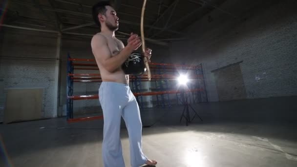Спортивный человек играет на национальном бразильском инструменте berimbau после выполнения капоэйра элементов в комнате с бетонным полом и кирпичными стенами — стоковое видео