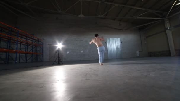 Un atlético haciendo elementos de capoeira y salta en la habitación con paredes de hormigón y ladrillo — Vídeos de Stock