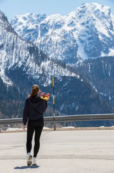 Une jeune femme debout sur un fond de montagnes et tenant un skateboard — Photo