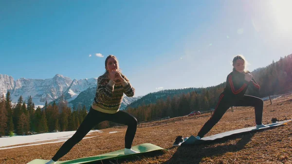 Dos mujeres jóvenes haciendo ejercicio al aire libre - en cuclillas y moverse a un lado - bosque y montañas en un fondo — Foto de Stock