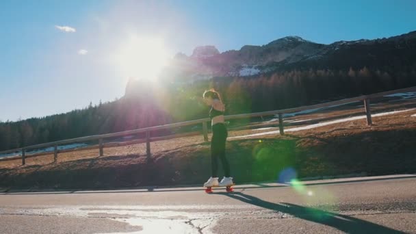 Mujer joven montando en la carretera en un pequeño monopatín en el fondo del bosque y las montañas — Vídeos de Stock