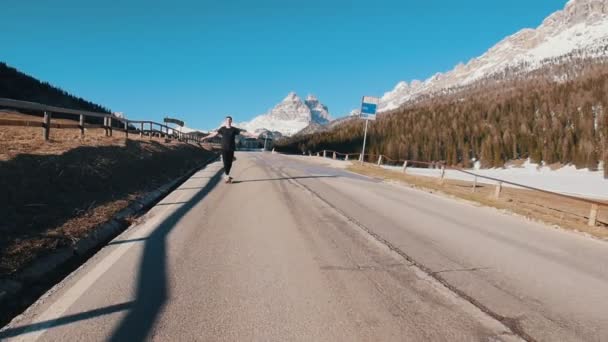 Молодой человек едет по дороге на небольшом скейтборде на фоне леса и гор — стоковое видео