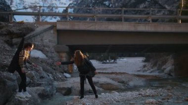 İki genç kadın dağlarda bir köprü altında sırt çantaları ve gitar ile kayalık bir nehir üzerinde gitmek