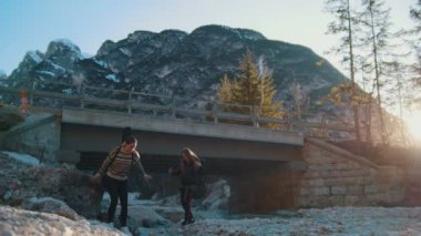 İki genç kadın dağlarda bir köprünün altında kayalık bir nehir üzerinde gitmek