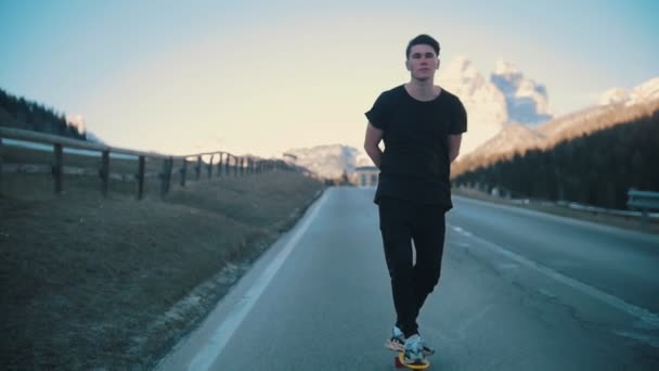 Молодой задумчивый человек едет по дороге с руками под спиной на маленьком скейтборде посреди Альп — стоковое видео