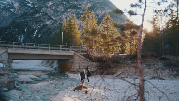 Dos mujeres jóvenes cruzando el río rocoso con las mochilas y la guitarra bajo un puente en las montañas — Vídeos de Stock