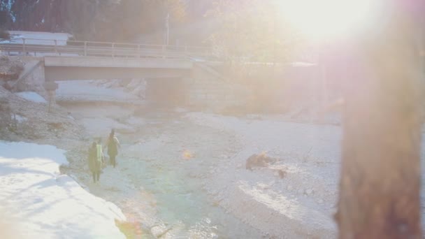 山の橋の下でバックパックとギターを持って岩の川の岸を歩く2人の若い女性 — ストック動画