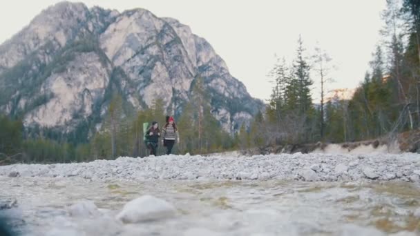 Две девушки, идущие вдоль берега скалистой реки с рюкзаками и гитарой на фоне леса и горы — стоковое видео
