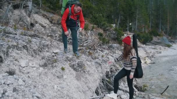 Ένας νέος άντρας με σακίδια που βοηθάνε τη γυναίκα με την κιθάρα να αναρριχηθεί στην ακτή του βραχώδους ποταμού στο φόντο του δάσους — Αρχείο Βίντεο