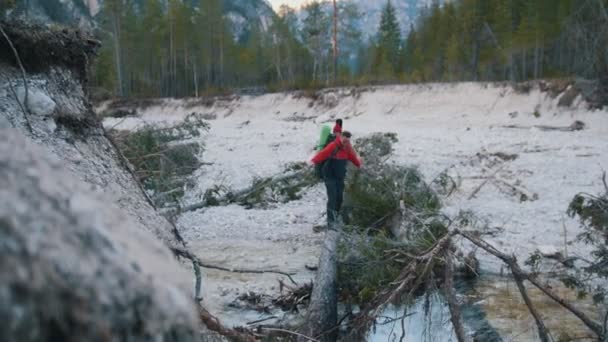 Een jonge man en vrouw gaan over de rotsachtige rivier op de log met rugzakken en gitaar op de achtergrond van het bos — Stockvideo