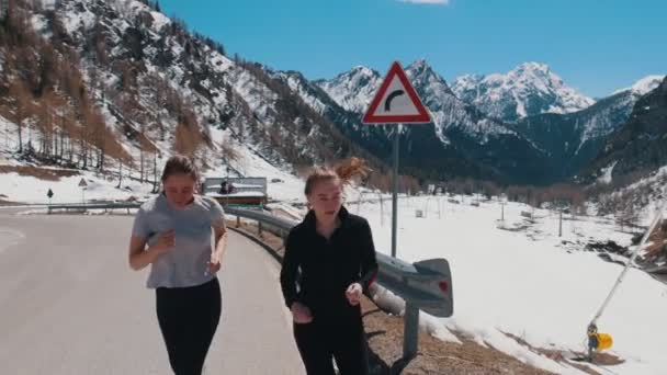 Zwei junge sportliche Frauen, die vor dem Hintergrund des schneebedeckten Berges die Straße hinunterlaufen — Stockvideo