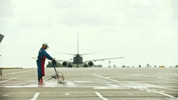 A man worker cleaning the asphalt on the airport field using water machine - a plane passing by — Stock Video