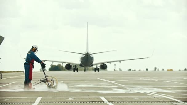 Um homem trabalhador limpando o solo no campo do aeroporto usando máquina de água - um avião que passa por — Vídeo de Stock