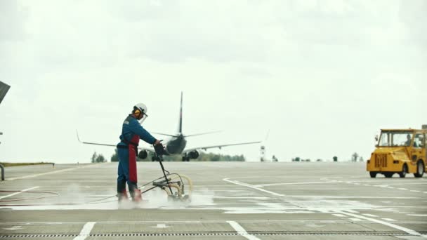 Un homme nettoie lentement l'asphalte sur le terrain de l'aéroport à l'aide d'une machine à eau - un avion passant par là — Video