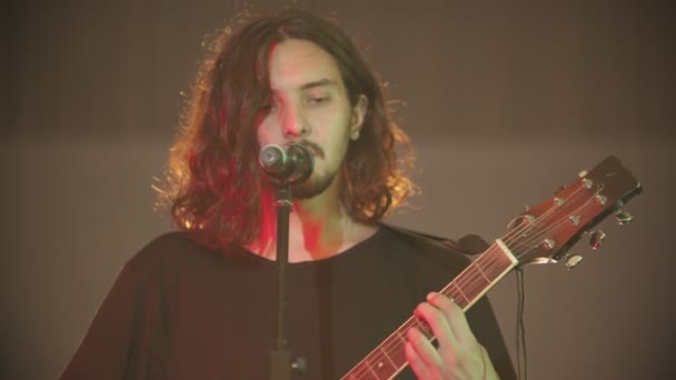 Молодий чоловік з довгим волоссям грає пісню на гітарі під час його виступу — стокове відео