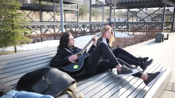 Jovem casal sentado no banco - um homem tocando guitarra — Vídeo de Stock