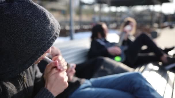 Junge Freunde chillen auf einer Bank - zwei Männer zünden sich Zigaretten an — Stockvideo