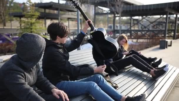 Młodzi przyjaciele schładza się na ławce-jeden facet bierze gitarę z innego i zaczyna grać — Wideo stockowe
