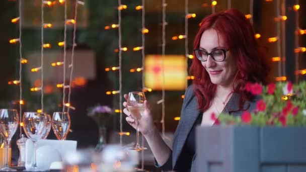 Деловая женщина с рыжими волосами сидит за столом в ресторане и держит бокал шампанского — стоковое видео