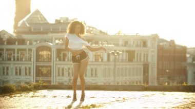 Küçük şortlu genç bir kadın çatıda çekici seksi dans yapıyor - squat oturur ve dans devam - parlak gün batımı