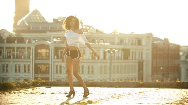 Una mujer joven en pantalones cortos pequeños de pie desde el suelo y realizando un atractivo baile sexy en el techo - puesta de sol brillante — Vídeos de Stock