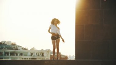 Kıvırcık saçlı ve küçük şortlu genç bir kadın çekici dans - aşağı gidiyor ve bacaklarını yayılıyor - gün batımı