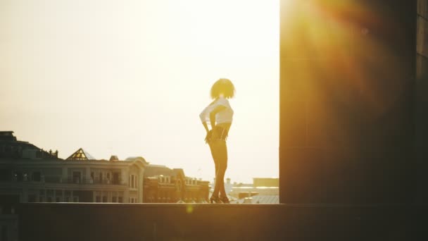Een jonge vrouw met krullend haar en in kleine shorts het uitvoeren van aantrekkelijke strip dansen op een achtergrond van gebouwen-zonsondergang — Stockvideo
