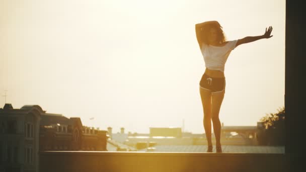 Uma jovem mulher atraente com cabelo encaracolado executando elementos de dança em um fundo de edifícios - pôr do sol — Vídeo de Stock