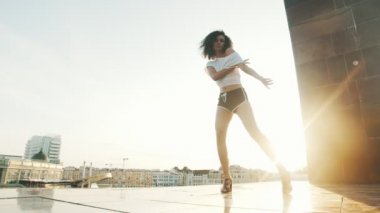 Kıvırcık saçlı genç bir kadın modern binaların arka planda çekici şerit dans performans - gün batımı