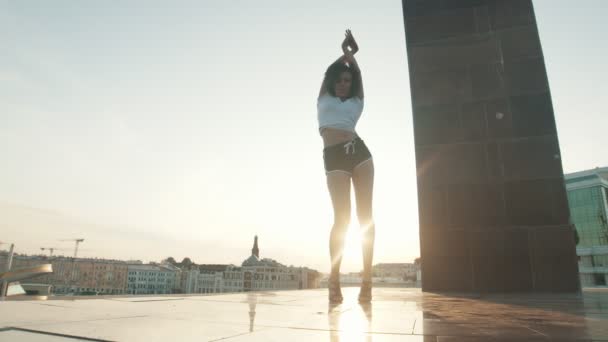 Молодая женщина смешанной расы с вьющимися волосами, исполняющая привлекательные танцы на фоне современных зданий - закат — стоковое видео