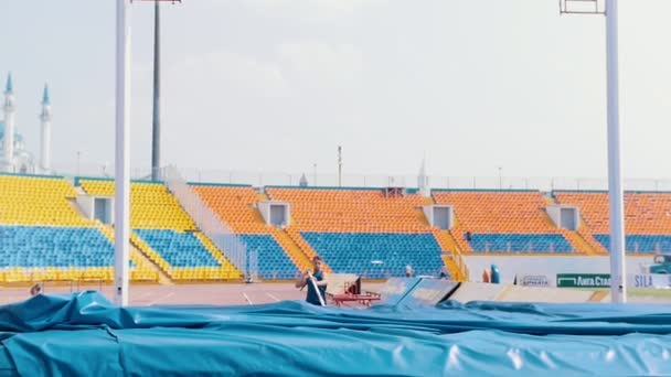 KAZAN, RÚSSIA 26-07-2019: um jovem de camisa azul correndo e se inclina em um poste para saltar sobre o bar - treinamento em salto com vara no estádio - tentativa mal sucedida por causa de tocar o bar — Vídeo de Stock