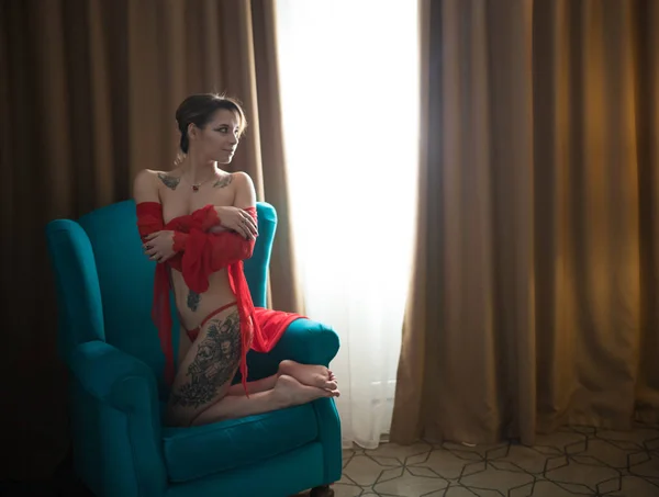 Jonge vrouw met tatoeages in een sexy rood ondergoed zittend in de stoel op haar knieën — Stockfoto