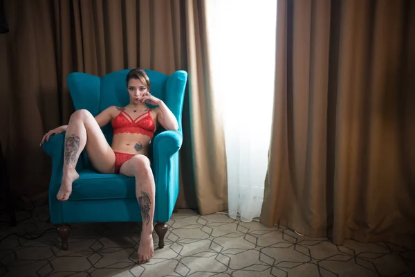 Молода впевнена жінка з татуюваннями в сексуальній червоній нижній білизні сидить у кріслі — стокове фото