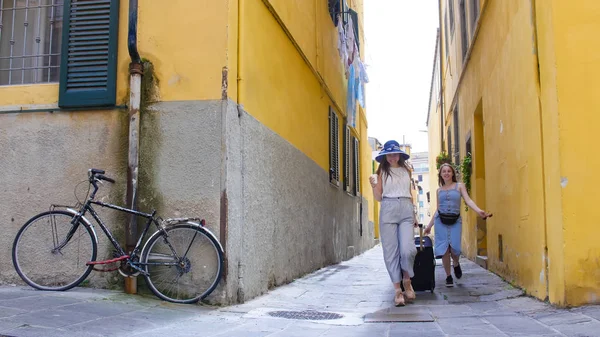 İki genç gülümseyen kadın bagaj ile dar sarı sokaklarda yürüyüş — Stok fotoğraf