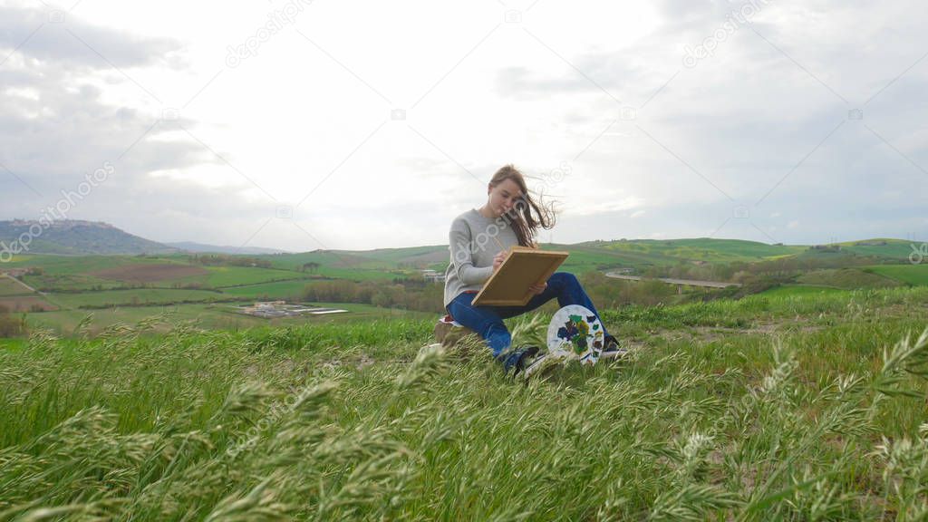 A young woman sits on the green field and drawing a painting on canvas - a palette in her legs