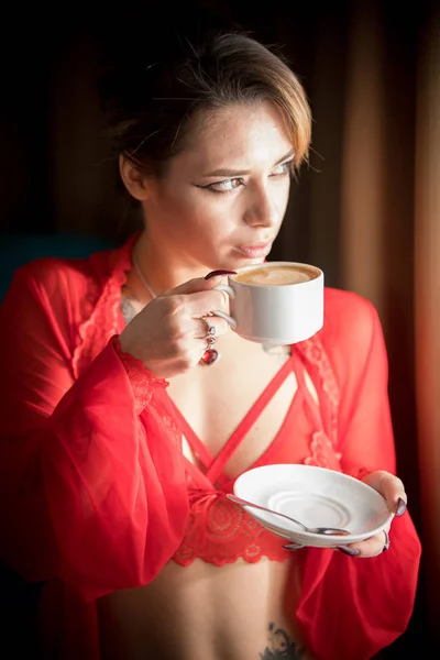 Jonge vrouw met tatoeages in een sexy rood ondergoed staande in de buurt van het raam en het drinken van een kopje koffie — Stockfoto