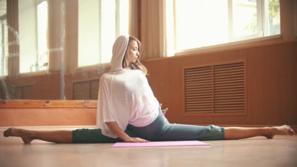 Beyaz gevşek gömlek genç kadın yoga mat üzerinde oturan bir bölünmüş performans - bacak germe egzersizleri yapıyor - dans stüdyosu — Stok video