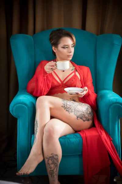 Mujer joven con tatuajes en ropa interior roja sexy sentada en una silla y bebiendo café —  Fotos de Stock