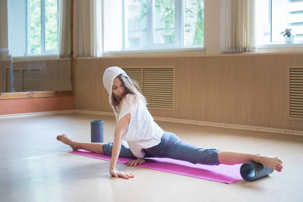 Junge schlanke Frau mit blonden Haaren sitzt auf der Yogamatte in Split - mit Hilfsständer unter dem Fuß - streckt ein Bein aus — Stockfoto