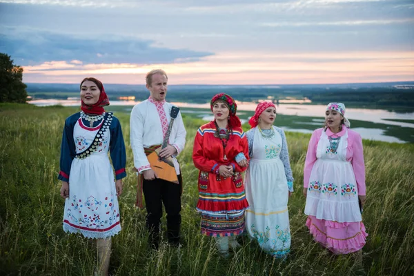 People in traditional russian clothes standing on a green field and singing - a man holding balalaika