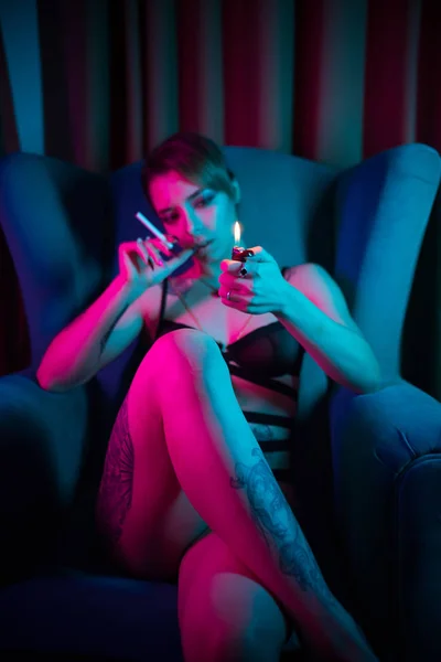 섹시한 속옷을 입은 젊은 여성이 다리를 교차하고 라이터를 들고 의자에 앉아 담배를 밝히려고합니다. — 스톡 사진