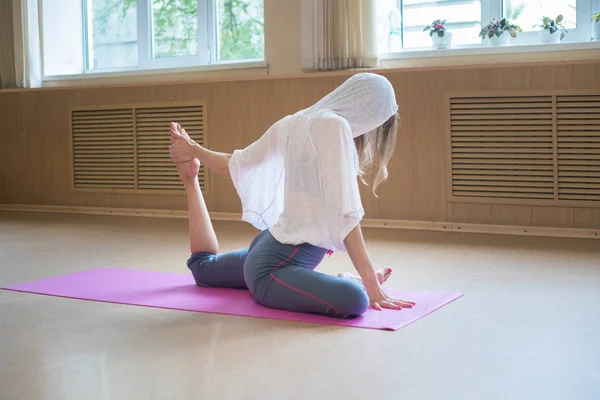 Joven mujer delgada con el pelo rubio sentado en la esterilla de yoga y haciendo ejercicios de estiramiento tira de la pierna hacia el cuerpo —  Fotos de Stock