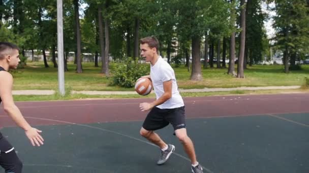 Deportista en camiseta blanca jugando baloncesto en la cancha al aire libre con un amigo, regateando y lanzando la pelota — Vídeo de stock