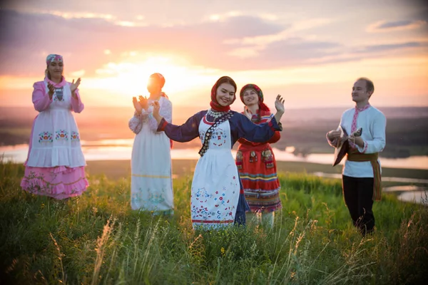 Люди в традиционной русской одежде стоят на поле на фоне яркого заката - танцует женщина — стоковое фото